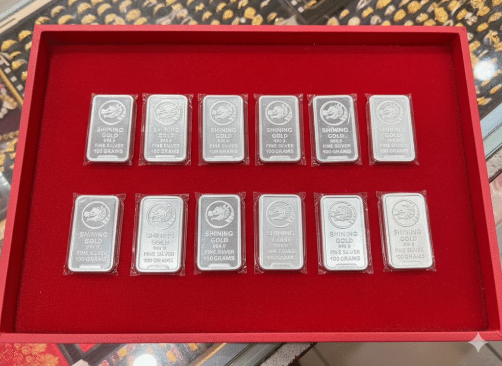 แท่งเงินสิบสองแท่ง แต่ละแท่งติดป้ายว่า "Shining Gold Fine Silver 100 กรัม" จัดเรียงเป็นสามแถวภายในตู้โชว์สีแดง เหมาะสำหรับนักสะสมหรือผู้ที่สนใจบริการตรวจสอบเปอร์เซ็นต์ทอง.