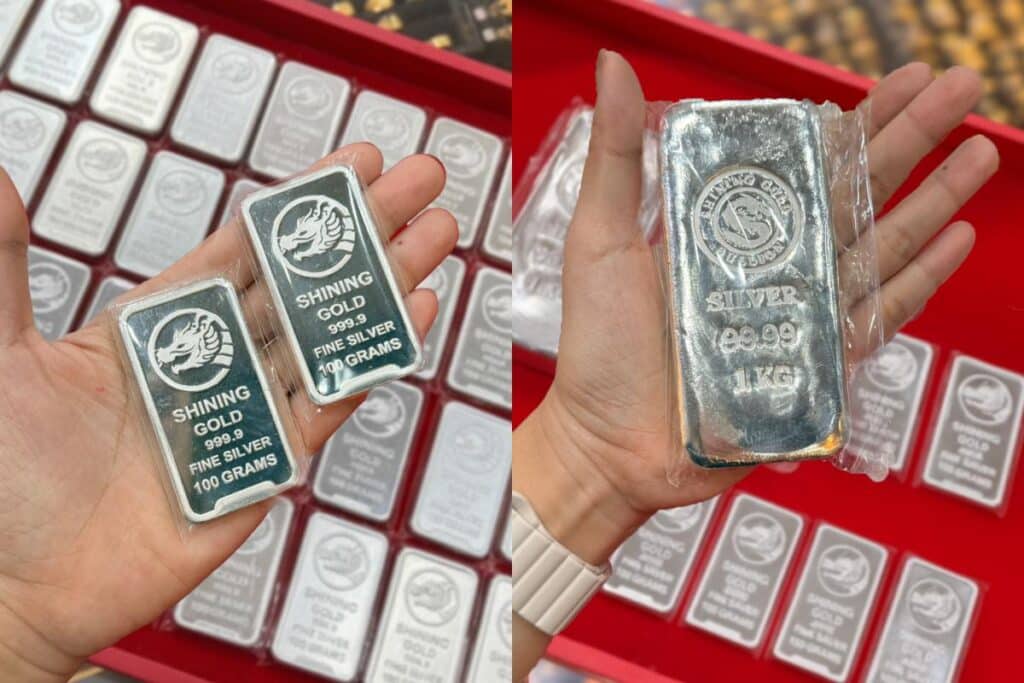 มือหนึ่งถือแท่งเงินสองแท่ง แท่งละ 100 กรัม และแท่งเงินหนึ่งแท่ง 1 กิโลกรัม; แท่งเงินที่มีฉลากคล้ายกันปรากฏอยู่ในพื้นหลัง เหมาะสำหรับนักสะสมหรือผู้ที่ต้องการตรวจสอบเปอร์เซ็นต์ทองคำ.