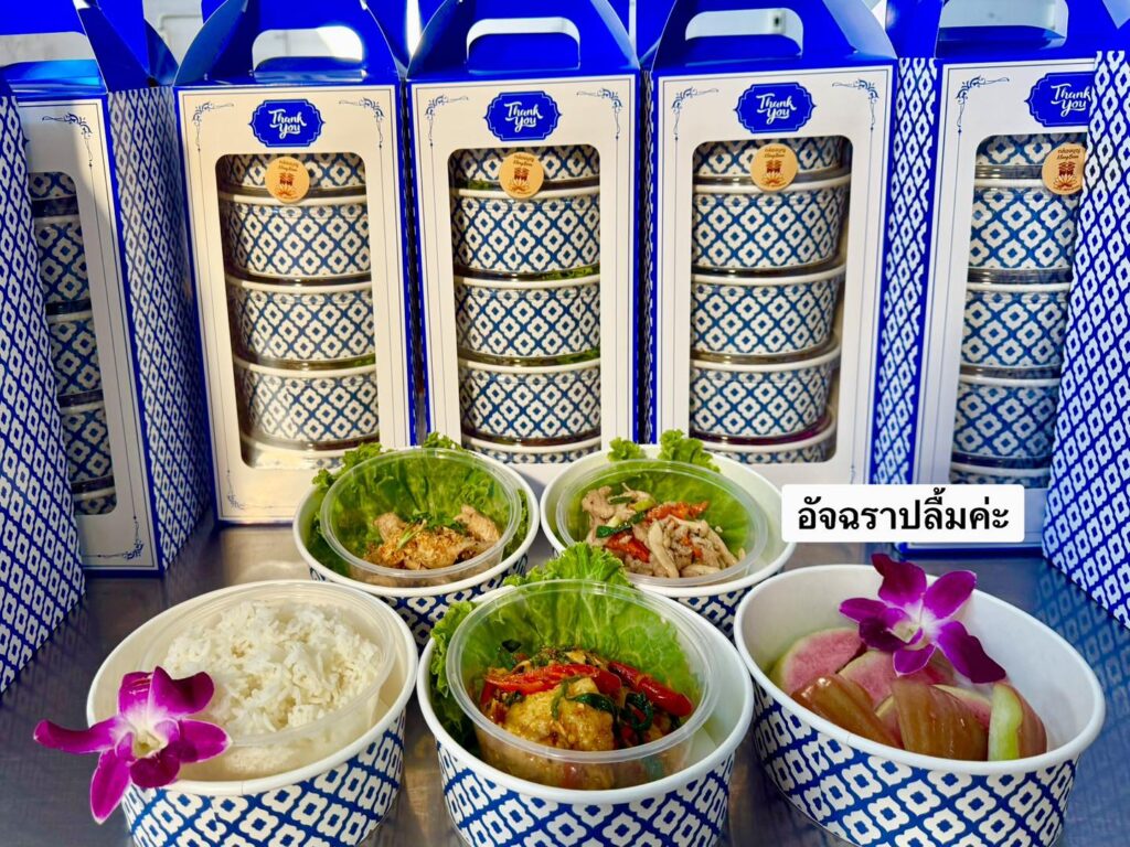 อาหารกล่องสไตล์ไทยในภาชนะลวดลายสวยงาม ประกอบด้วยอาหารสี่อย่างตกแต่งด้วยดอกไม้และผักกาดหอม จัดวางอยู่หน้ากล่องอาหารแบบซ้อนกัน มีฉลากภาษาไทยแนบมาด้วย ให้ความรู้สึกหรูหราคล้ายบรรจุภัณฑ์ที่พบใน ร้านทองออนไลน์.