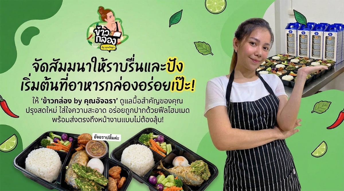 ผู้หญิงคนหนึ่งยืนอยู่ข้างชุดอาหารบรรจุกล่องบนโต๊ะ หน้าป้ายโฆษณาออนไลน์ร้านทองที่มีสีสัน ซึ่งโฆษณาอาหารกล่องพร้อมรับประทานในภาษาไทย.