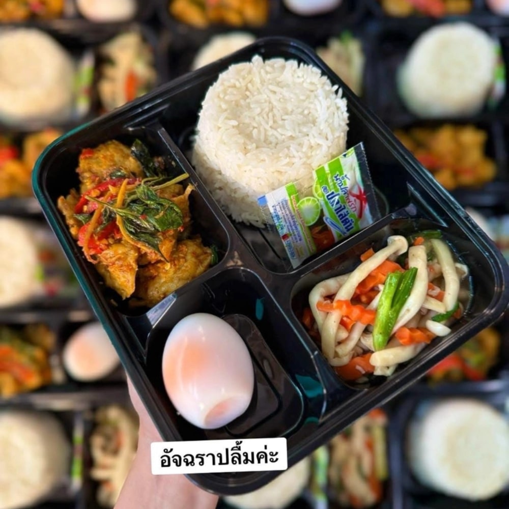 มือหนึ่งถือถาดอาหารสีดำที่มีข้าวสวย ไข่ต้ม ผัดผัก อาหารแกง และซองเครื่องปรุงรสเล็กๆ หนึ่งซอง—ชวนให้นึกถึงมื้ออาหารที่คุณอาจเพลิดเพลินขณะเลือกชม ร้านทองออนไลน์ ถาดอื่นๆ ปรากฏเป็นภาพเบลออยู่เบื้องหลัง.