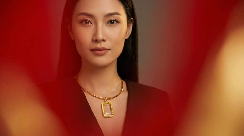 ผู้หญิงคนหนึ่งมีผมยาวสีดำ สวมสร้อยคอทองคำซึ่งอาจมีจี้กรอบพระประดับอยู่ และสวมชุดสีเข้ม มองตรงไปที่กล้องด้วยสีหน้าเรียบเฉย รูปทรงสีแดงและสีเหลืองล้อมรอบภาพ.
