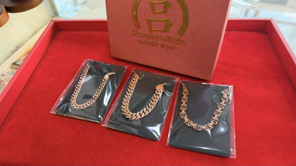 สร้อยข้อมือทองสามเส้นที่จัดแสดงในบรรจุภัณฑ์สีดำ วางบนพื้นผิวกำมะหยี่สีแดง ข้างกล่องสีแดงที่มีข้อความและโลโก้ภาษาไทยสีทอง เหมาะสำหรับผู้ที่สนใจ ราคานาควันนี้ 2569 ล่าสุด บาทละ หรือกำลังมองหา สร้อยนาค ที่มีสไตล์เพื่อเสริมความสมบูรณ์ให้คอลเลกชันใดๆ.